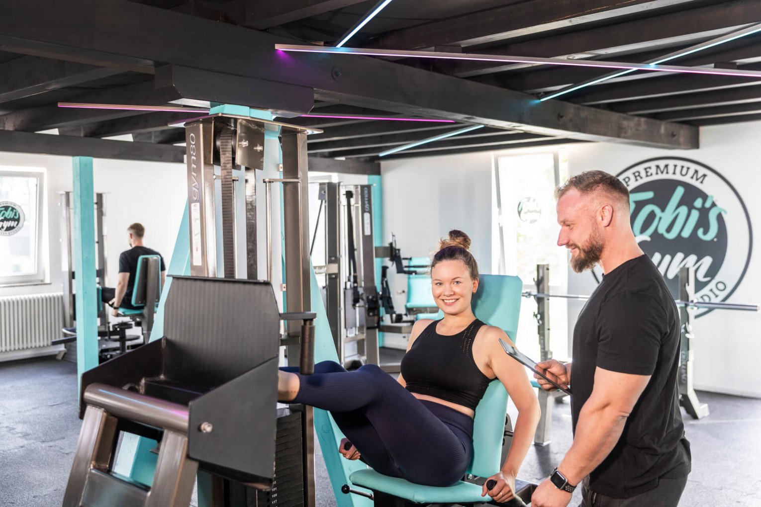 Sportlerin trainiert auf der Beinpresse in Tobi's Gym, Trainer steht daneben